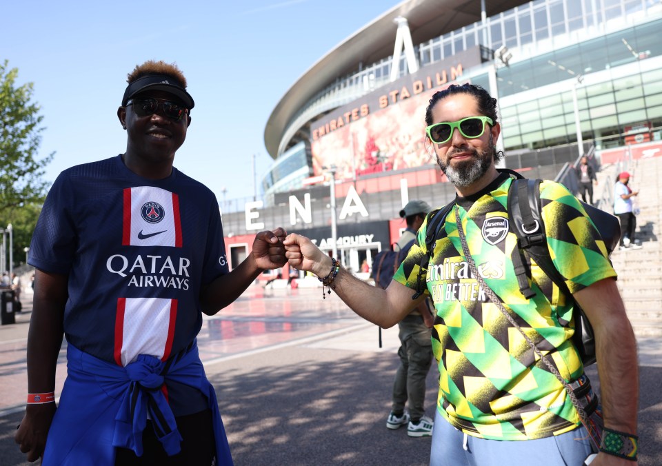 Two soccer fans, one wearing a Paris Saint-Germain jersey and the other an Arsenal jersey, bump fists outside the Emirates Stadium.